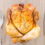 Top view of a perfectly roasted chicken placed between fork and knife on a wooden table.