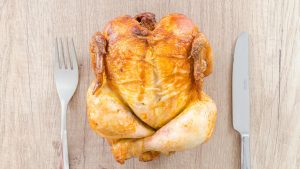 Top view of a perfectly roasted chicken placed between fork and knife on a wooden table.