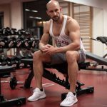 Strong, determined man in white tank top sitting in gym, showcasing fitness and strength.